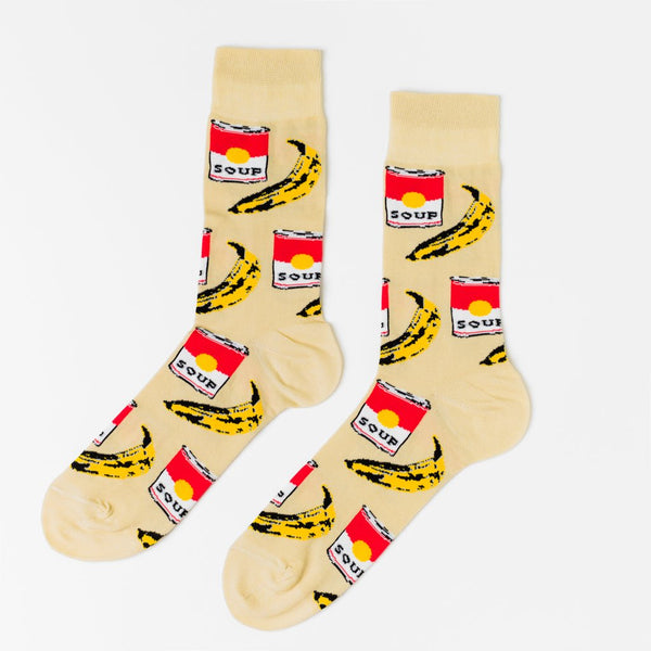 Shown in a relaxed flatlay, a pair of Yellow Owl Workshop’s tan nylon-cotton men's crew socks with browning bananas and red soup cans