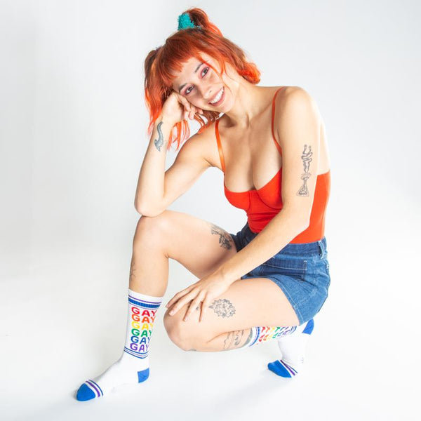A model wearing white cotton unisex crew socks with a blue striped toe and cuff by the brand Gumball Poodle feature the word "GAY" repeated down the leg in rainbow colors.