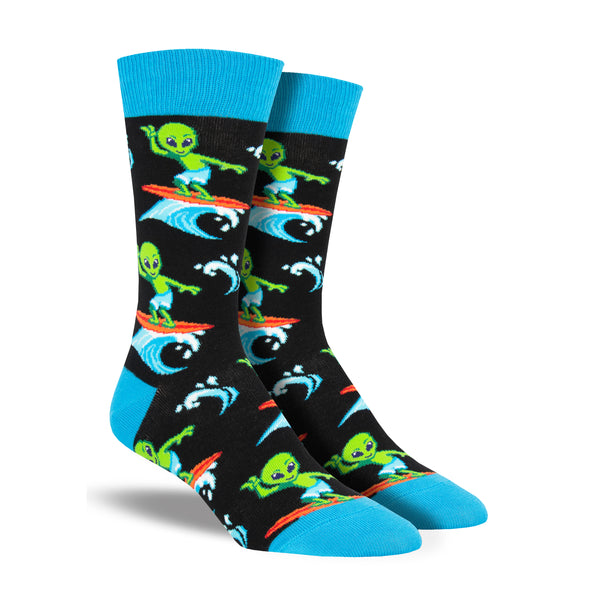Shown on a foot form, a pair of Socksmith's black cotton men's crew socks with blue heel/cuff/toe and all-over pattern of a goofy, green alien surfing a blue ocean wave