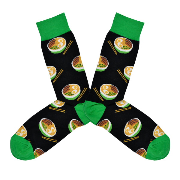 Shown in a flatlay, a pair of Socksmith brand men's cotton crew socks in black with a green cuff/heel/toe featuring an all over motif of bowls of ramen and chopsticks.