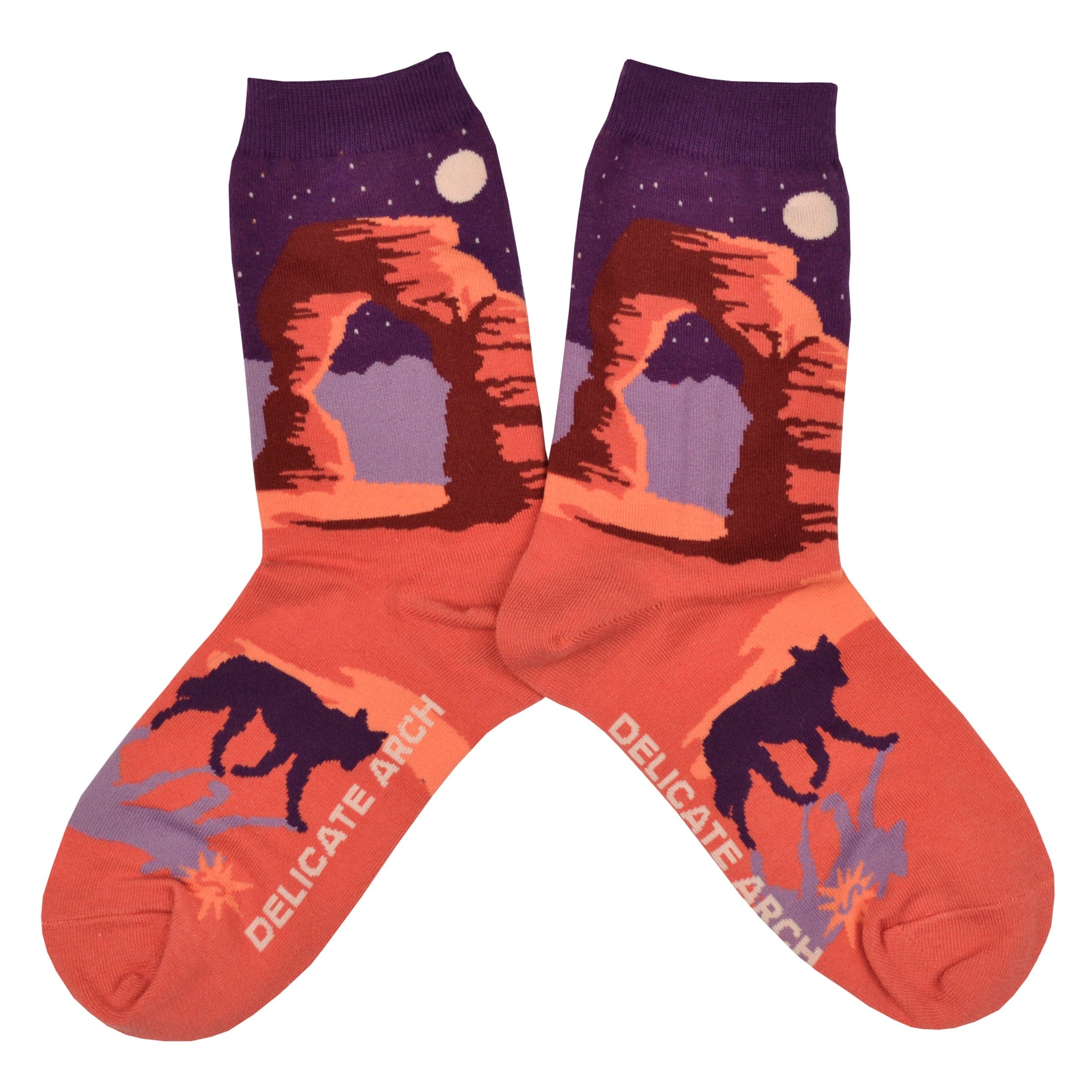 Shown in a flatlay, a pair of Sock It to Me brand women's cotton crew socks with an orange heel/toe an a purple cuff. These socks feature an arch from Arches National Park on the leg and a coyote on the foot of the sock as well as the words 