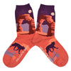 Shown in a flatlay, a pair of Sock It to Me brand women's cotton crew socks with an orange heel/toe an a purple cuff. These socks feature an arch from Arches National Park on the leg and a coyote on the foot of the sock as well as the words "delicate arch" along the foot.