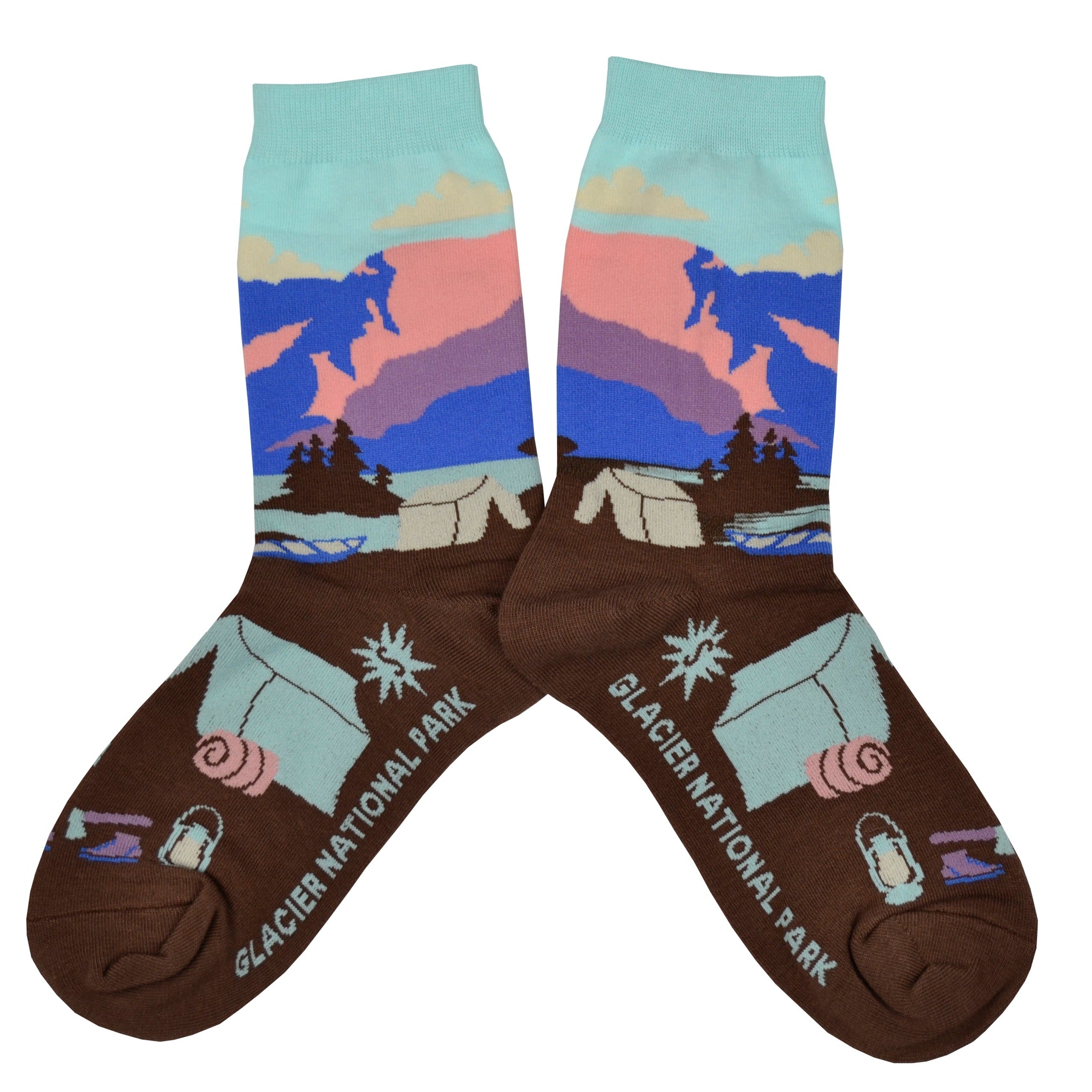 Shown in a flatlay, a pair of women's Sock it to Me cotton crew socks with a sea foam green cuff and a brown heel and toe. These socks feature a depiction of Montana's Glacier National Park with a sea foam green, pink, and blue leg with brown trees and a brown foot with a camping scene.
