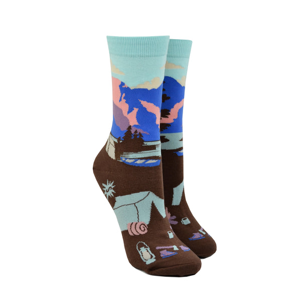 Shown on leg forms, a pair of women's Sock it to Me cotton crew socks with a sea foam green cuff and a brown heel and toe. These socks feature a depiction of Montana's Glacier National Park with a sea foam green, pink, and blue leg with brown trees and a brown foot with a camping scene.