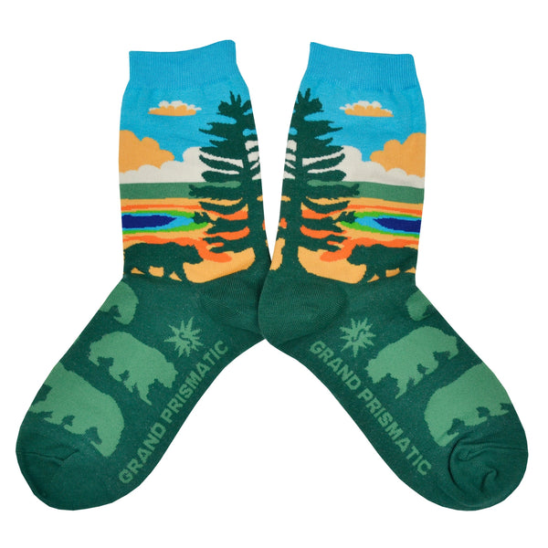 Shown in a flatlay, a pair of women's Sock it to Me cotton crew socks with the Yellowstone National Park Grand Prismatic Spring depicted on the sock. The leg features the spring with a blue sky background while the foot is dark green with light green bear silhouettes.