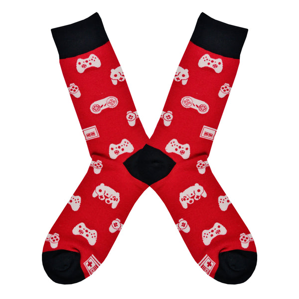 Shown in a flatlay, a pair of Sock it to Me brand men's cotton crew socks in red with a black heel, toe, and cuff. These socks feature various vintage game controllers in white all over the sock.