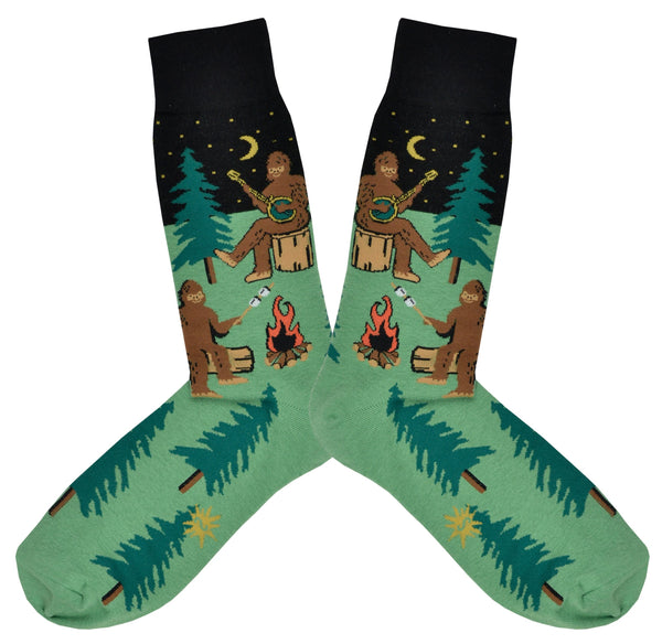 Shown in a flatlay, a pair of Sock It To Me green cotton men's crew socks with black starry sky and various sasquatches enjoying a campfire (playing banjo, roasting marshmallows) among a pine forest
