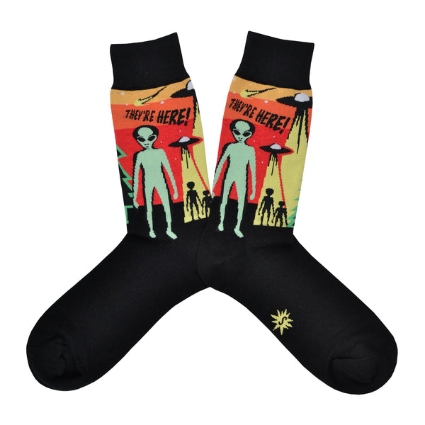 Shown in a flatlay, a pair Sock It to Me cotton men's crew socks in black with a yellow, orange, and red background on the leg featuring a green alien and 2 space ships. The text on the sock reads, "They're Here!".