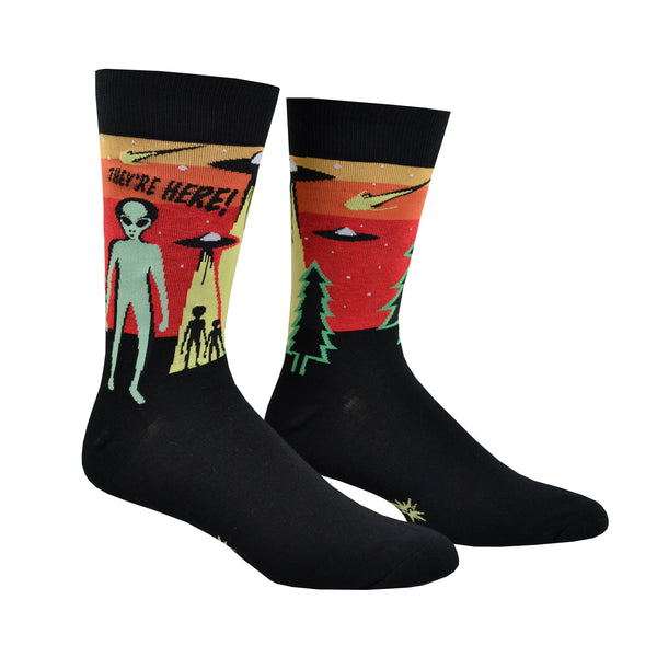 Shown on foot forms, a pair Sock It to Me cotton men's crew socks in black with a yellow, orange, and red background on the leg featuring a green alien and 2 space ships. The text on the sock reads, "They're Here!".