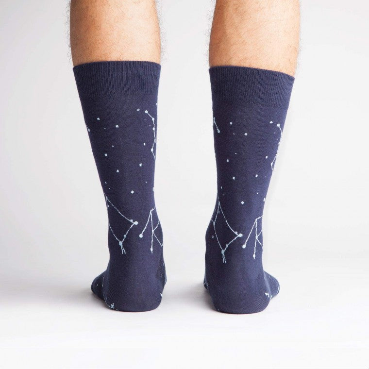 Shown on a model, a pair of Sock It To Me navy blue cotton men's crew socks with glow in the dark constellation all over pattern