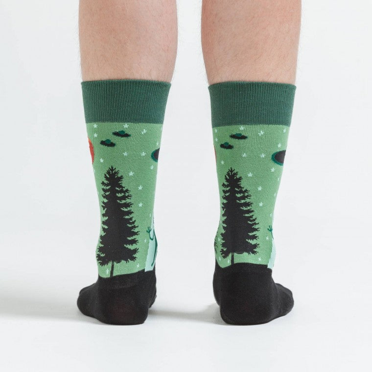 Shown on a model, a pair of Sock It To Me green cotton men's crew socks with alien waving goodbye as it gets sucked into a UFO