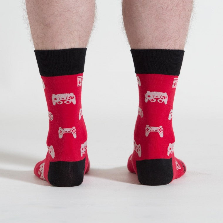 Shown from the back on a light skinned model, a pair of Sock it to Me brand men's cotton crew socks in red with a black heel, toe, and cuff. These socks feature various vintage game controllers in white all over the sock.