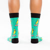 Shown on a model, a pair of Sock It To Me turquoise cotton men's crew socks with black cuff/heel/toe and goofy bananas skateboarding while wearing cool sunglasses
