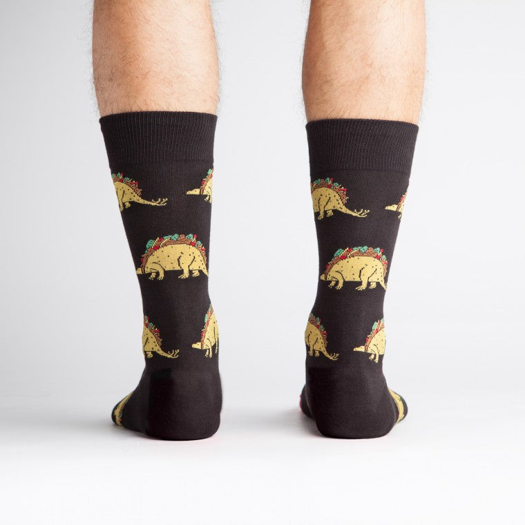 A model wearing black cotton men's crew socks by the brand Sock It To Me featuring dinosaurs with bodies that look like a taco.