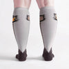 Shown from the back on a light skinned model with larger calves, a pair of Sock It To Me brand Stretch It cotton knee high socks in grey and black. The leg of the sock is grey with a cartoon ostrich design going down the leg and transitioning into the black foot of the sock. This image displays the Stretch It technology that allows this sock to stretch up to 21 inches around the leg.