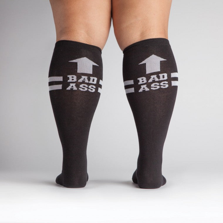 Shown from the back a dark skinned model with wide calves wears a pair of Sock It To Me brand unisex cotton knee high socks in black with two white stripes around the calf. the back of the sock features a white arrow pointing up to the wearer and says, 