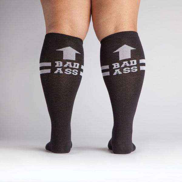 Shown from the back a dark skinned model with wide calves wears a pair of Sock It To Me brand unisex cotton knee high socks in black with two white stripes around the calf. the back of the sock features a white arrow pointing up to the wearer and says, "BAD ASS".