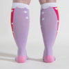 Shown on a model with large calves, a pair of Sock it to Me brand unisex cotton knee high socks in lilac with a pink heel, cuff, and toe. The sides of this sock feature a cartoon unicorn farting a magical rainbow that goes up the sock. This image shows just how much the Stretch-It technology allows these socks to stretch without distorting the image.