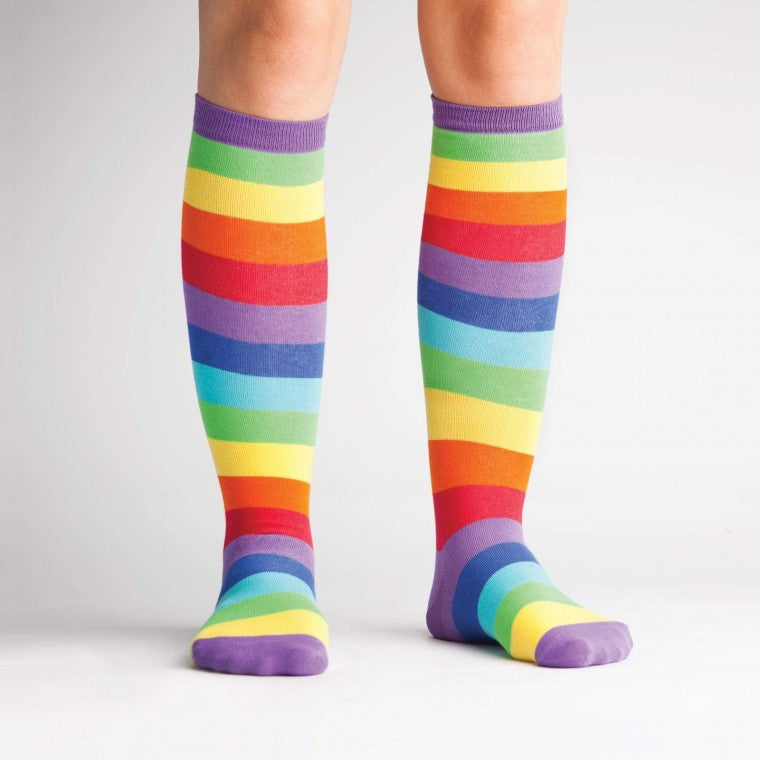 Shown on a light-skinned model, a pair of Sock it to Me brand Stretch-It cotton knee high socks with an all around rainbow stripe. The heel, toe, and cuff are done in purple while the red of the sock is classic ROYGBIV rainbow.