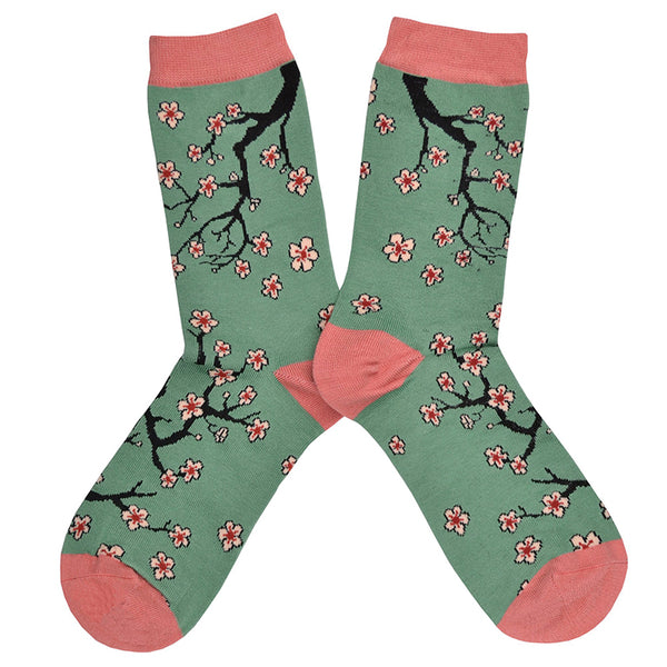 Shown in a flatlay, a pair of Socksmith brand women's bamboo crew socks in sage green with a pink heel, toe, and cuff. This sock features an all over design cherry blossom branches reaching around the sock.