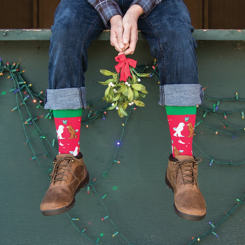 A pair of legs dangle from a stoop in rolled up denim jeans, brown boots, and the Men's Mythical Kissmas socks, the model holds a mistletoe between their knees.