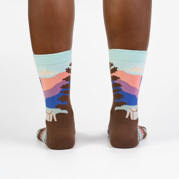 Shown on a dark skinned model from the back, a pair of women's Sock it to Me cotton crew socks with a sea foam green cuff and a brown heel and toe. These socks feature a depiction of Montana's Glacier National Park with a sea foam green, pink, and blue leg with brown trees and a brown foot with a camping scene.