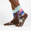 Shown on a dark skinned model from the side, a pair of women's Sock it to Me cotton crew socks with a sea foam green cuff and a brown heel and toe. These socks feature a depiction of Montana's Glacier National Park with a sea foam green, pink, and blue leg with brown trees and a brown foot with a camping scene.