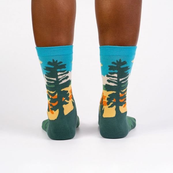 Shown from the back on a dark skinned model, a pair of women's Sock it to Me cotton crew socks with the Yellowstone National Park Grand Prismatic Spring depicted on the sock. The leg features the spring with a blue sky background while the foot is dark green with light green bear silhouettes.