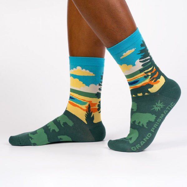 Shown on a dark skinned model's feet, a pair of women's Sock it to Me cotton crew socks with the Yellowstone National Park Grand Prismatic Spring depicted on the sock. The leg features the spring with a blue sky background while the foot is dark green with light green bear silhouettes.