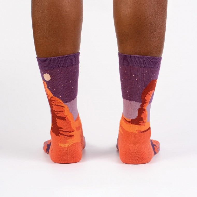 Shown on a dark skinned model from the back, a pair of Sock It to Me brand women's cotton crew socks with an orange heel/toe an a purple cuff. These socks feature an arch from Arches National Park on the leg and a coyote on the foot of the sock as well as the words 