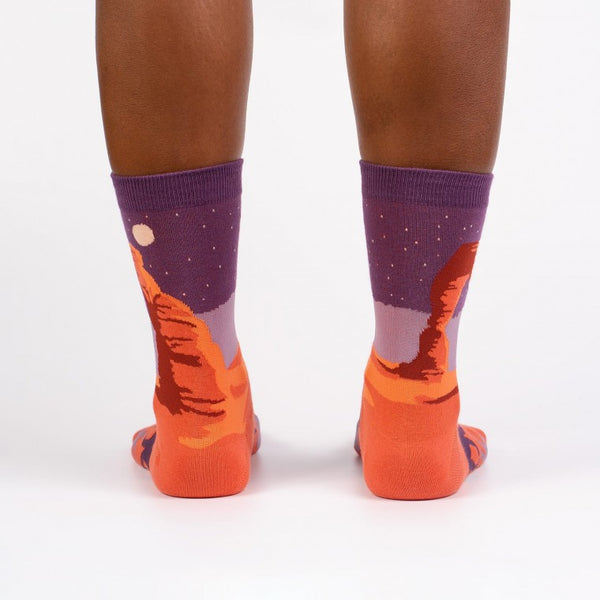 Shown on a dark skinned model from the back, a pair of Sock It to Me brand women's cotton crew socks with an orange heel/toe an a purple cuff. These socks feature an arch from Arches National Park on the leg and a coyote on the foot of the sock as well as the words "delicate arch" along the foot.