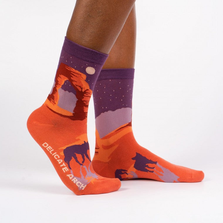 Shown on a dark skinned model with one foot cocked, a pair of Sock It to Me brand women's cotton crew socks with an orange heel/toe an a purple cuff. These socks feature an arch from Arches National Park on the leg and a coyote on the foot of the sock as well as the words 