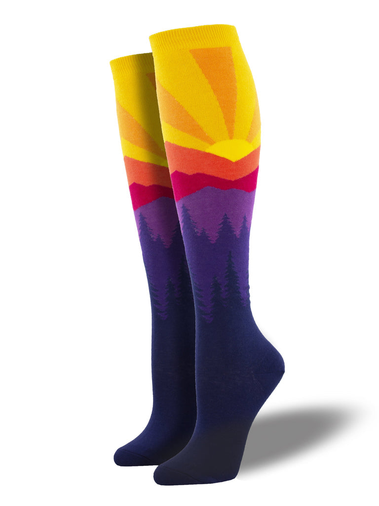 knee high socks on leg forms with a dark purple foot, lighter purple silhouettes of pine trees, purple, red and orange mountains on the horizon topped by a brilliant yellow sun with orange sunrays near the cuff