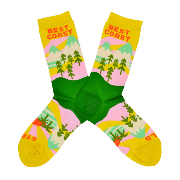 These yellow cotton women's novelty crew socks by the brand Yellow Owl Workshop feature green trees, teal and white mountains, and pink clouds and say the words "West Coast Best Coast" near the cuff and "Stay Gold" by the toes.