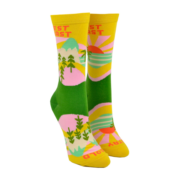 Shown on a leg form, these yellow cotton women's novelty crew socks by the brand Yellow Owl Workshop feature green trees, teal and white mountains, and pink clouds and say the words "West Coast Best Coast" near the cuff and "Stay Gold" by the toes.
