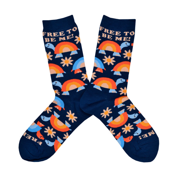Shown in a flatlay, a pair of women's Yellow Owl Workshop brand cotton and nylon socks in navy blue with orange and blue rainbow shelled turtles and orange daisies and the words "Free To Be Me" along the cuff.