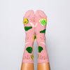 Shown on a model's feet, a pair of Yellow Owl Workshop brand women's cotton crew socks in a light brown with an avocado toast theme. Each sock features avocado toast, avocados, salt and pepper, and a knife.