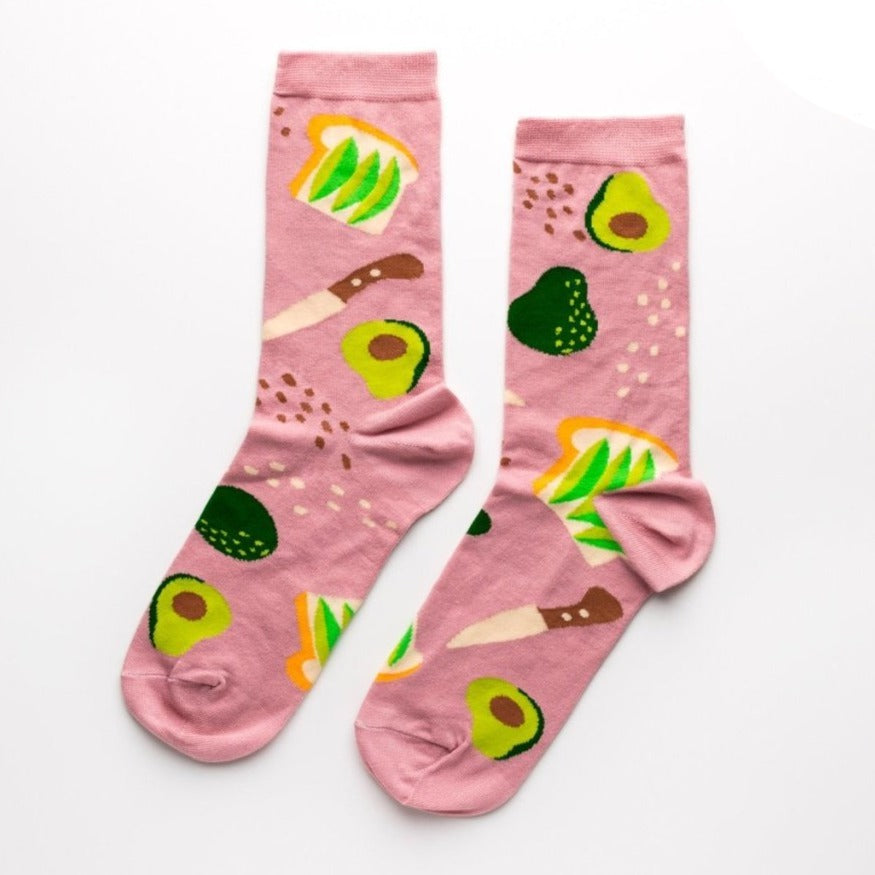 Shown in a front/back flatlay, a pair of Yellow Owl Workshop brand women's cotton crew socks in a light brown with an avocado toast theme. Each sock features avocado toast, avocados, salt and pepper, and a knife.