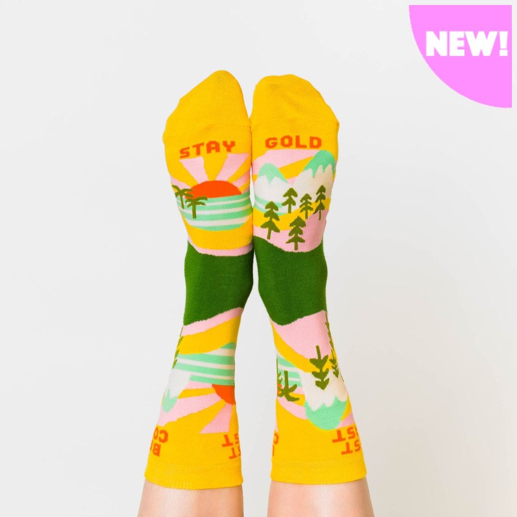 A model wearing yellow cotton women's novelty crew socks by the brand Yellow Owl Workshop feature green trees, teal and white mountains, and pink clouds and say the words 