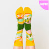 A model wearing yellow cotton women's novelty crew socks by the brand Yellow Owl Workshop feature green trees, teal and white mountains, and pink clouds and say the words "West Coast Best Coast" near the cuff and "Stay Gold" by the toes.