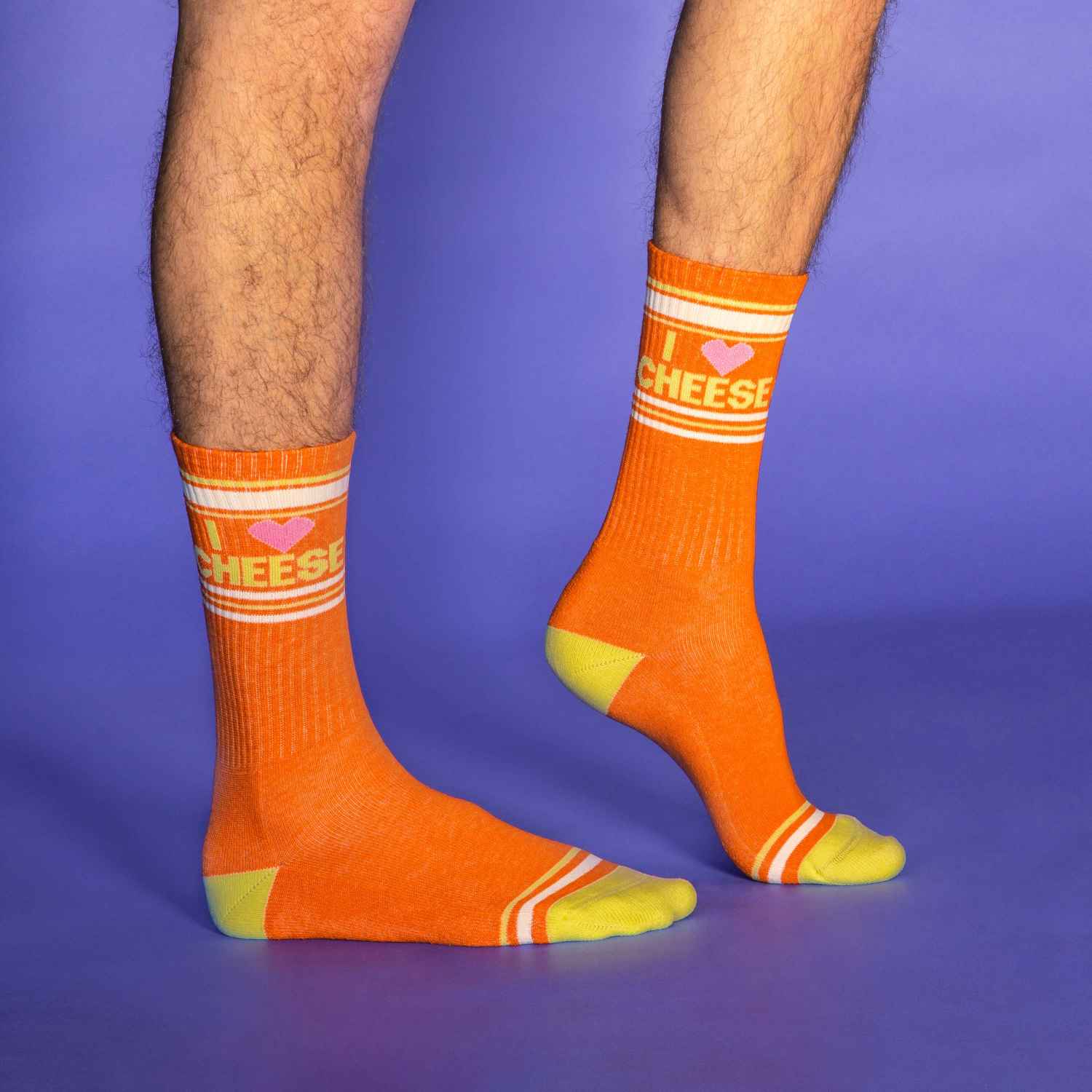 A purple background, a person's feet and lower legs. The feet are clad in orange crew socks with yellow heels, yellow & off-white stripe patterns near the toe and ankle, and yellow text that states 