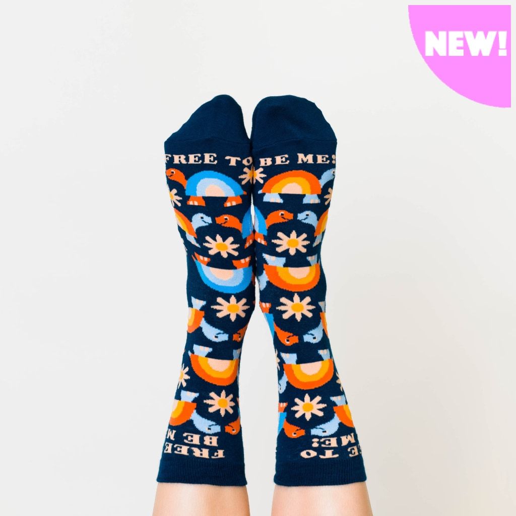 Shown on a models feet from the top, a pair of women's Yellow Owl Workshop brand cotton and nylon socks in navy blue with orange and blue rainbow shelled turtles and orange daisies and the words 