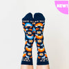 Shown on a models feet from the top, a pair of women's Yellow Owl Workshop brand cotton and nylon socks in navy blue with orange and blue rainbow shelled turtles and orange daisies and the words "Free To Be Me" along the cuff.