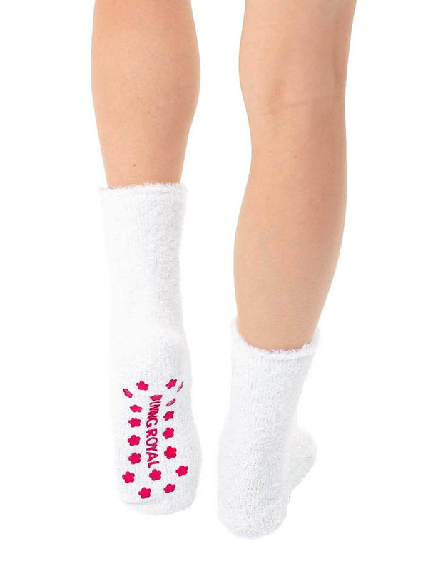 a pair of legs wearing a white fuzzy pair of crew length socks seen from the back with one foot raised so you can see the pink star shaped non skid grips on the sole