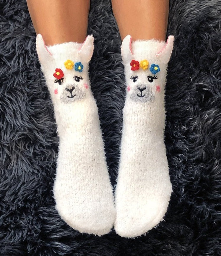  a pair of feet wearing fuzzy white socks with 3 dimensional ears and a face of a llama, decorated with small 3 dimensional flowers around the head of the llama on a grey furry background