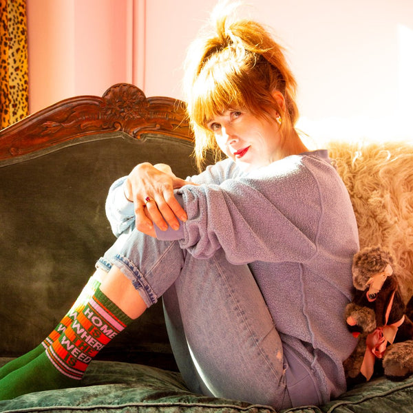 A model wearing green cotton unisex crew socks with an orange and pink striped toe and cuff by the brand Gumball Poodle feature the words "HOME IS WHERE THE WEED IS" on the leg.