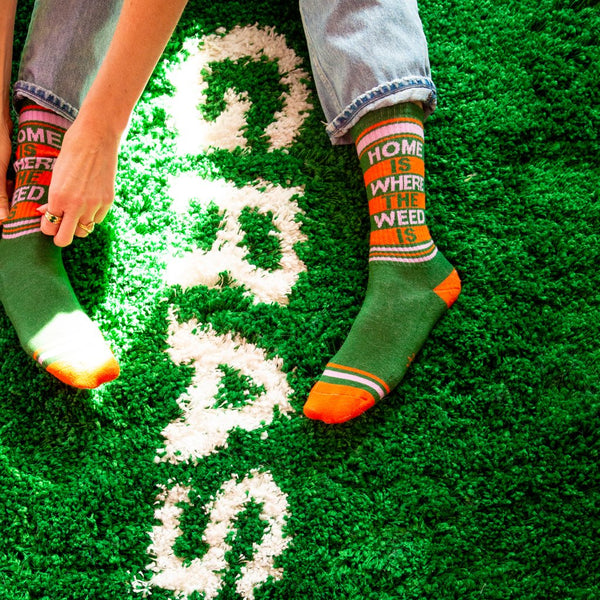A model wearing green cotton unisex crew socks with an orange and pink striped toe and cuff by the brand Gumball Poodle feature the words "HOME IS WHERE THE WEED IS" on the leg while sitting on a carpet that says "GRASS".