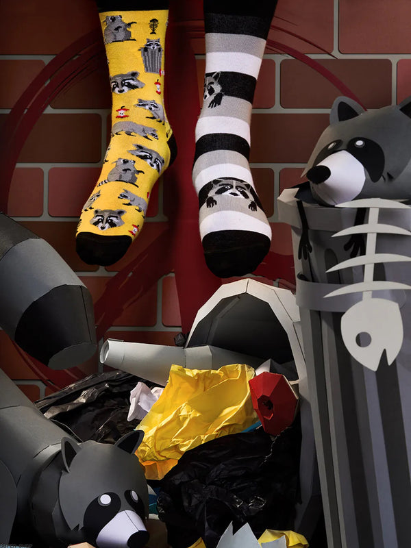 a pair of feet wearing mismatched yellow and gray raccoon print socks in front of a brick wall, surrounded by paper art depicting raccoons, trash cans and trash