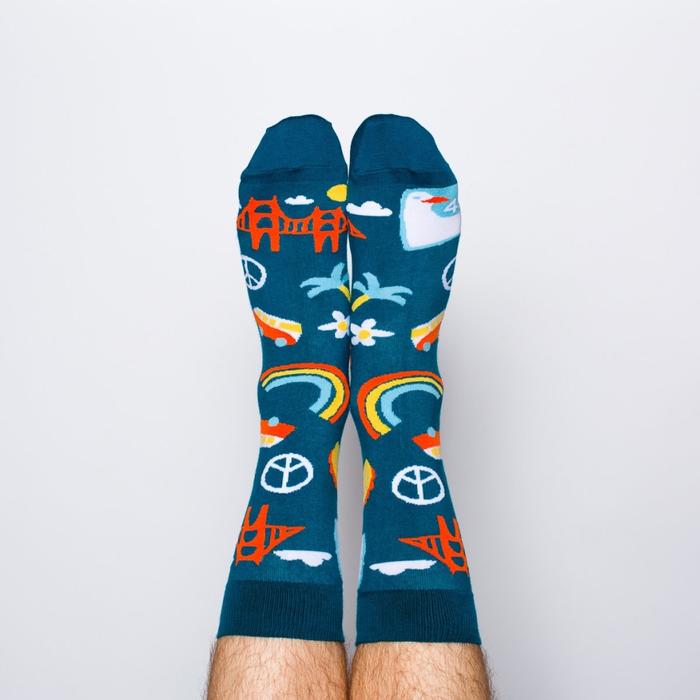 Model wearing a pair of Yellow Owl Workshop’s navy blue nylon-cotton men's crew socks with San Francisco Golden Gate Bridge, peace sign, palm tree, daisy, VW bus and highway One sign