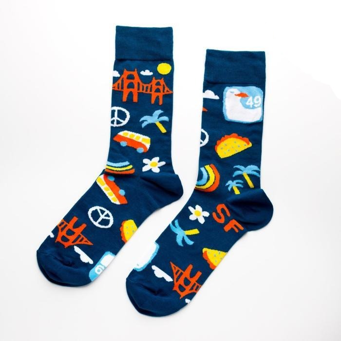 Shown in a relaxed flatlay, a pair of Yellow Owl Workshop’s navy blue nylon-cotton men's crew socks with San Francisco Golden Gate Bridge, peace sign, palm tree, daisy, VW bus and highway One sign